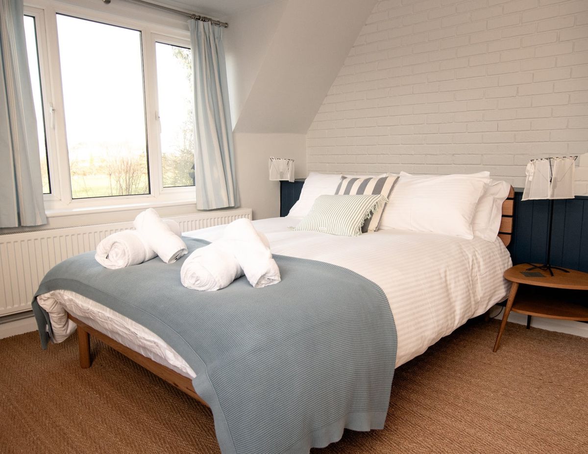 A bedroom under a sloping ceiling with a large bed in white linen, a grey throw, striped cushions and two windows