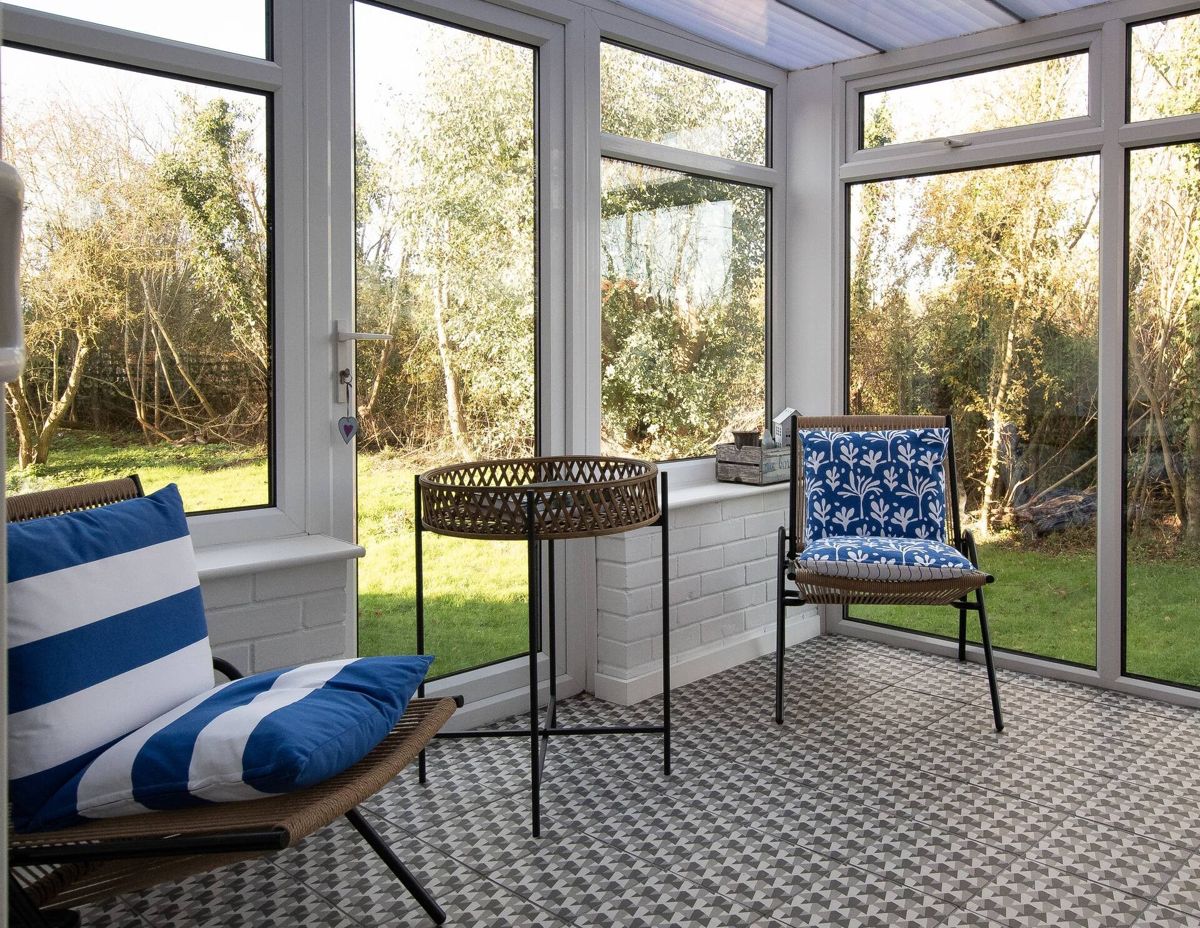 A conservatory with blue and white striped chairs, a small round table and glass walls looking out to trees