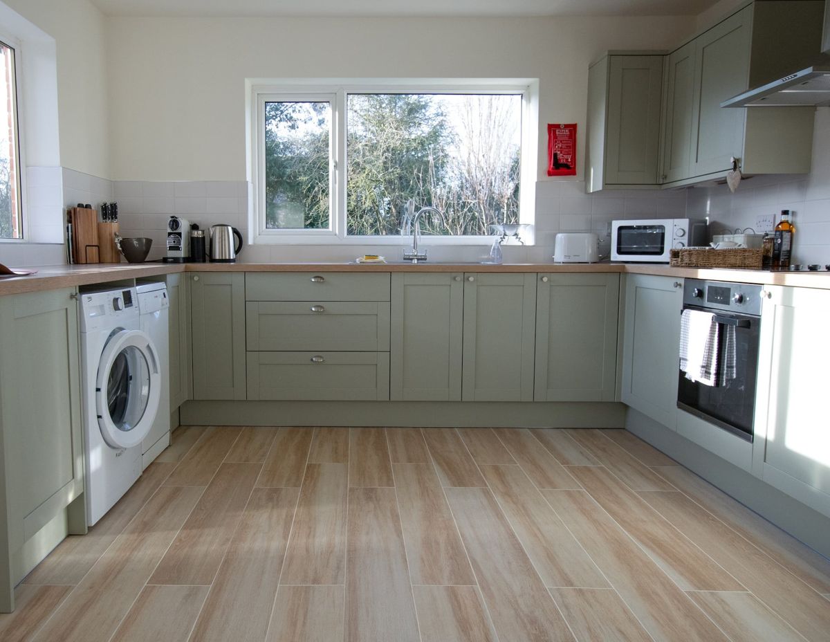 A long kitchen with sage green units, a washing machine, an oven and a window at the far end