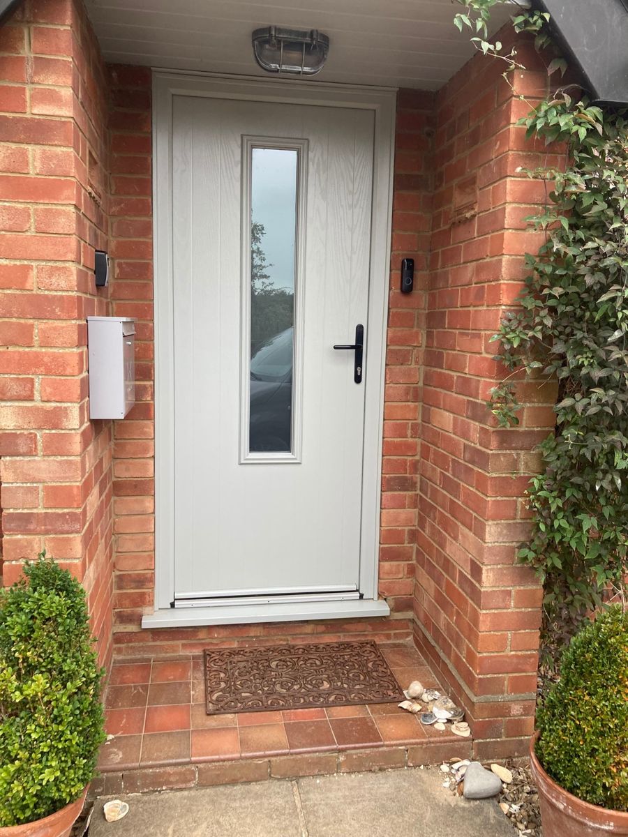 A grey front door with a glass panel in a red-brick porch, with clipped plants in pots either side